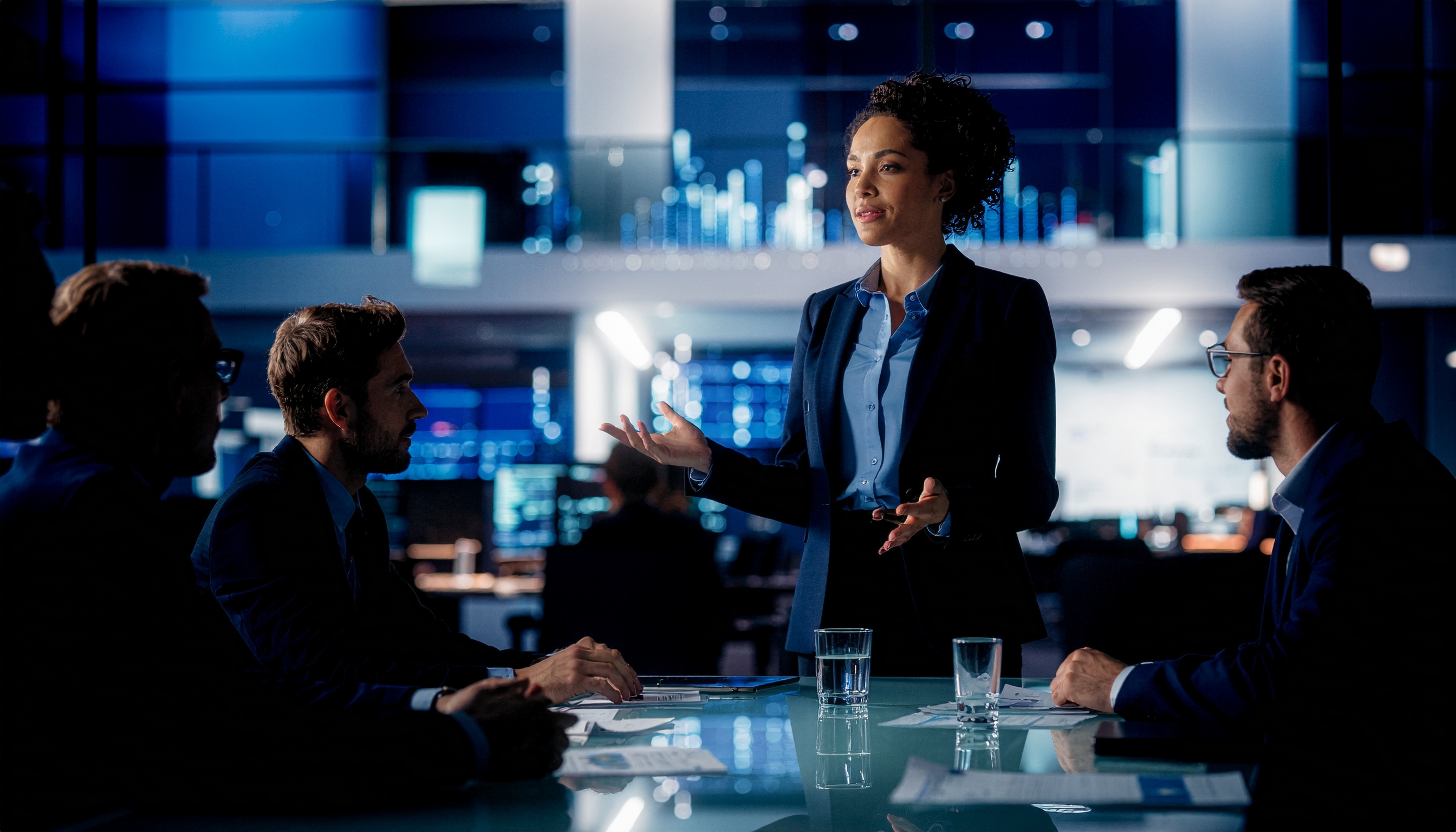 Cena corporativa noturna em uma sala de reuniões moderna, com uma mulher em pé apresentando, cercada por três homens sentados. A iluminação azul suave e as telas digitais ao fundo criam uma atmosfera tecnológica e profissional. Os detalhes dos ternos escuros e a mesa de vidro conferem um toque de sofisticação e foco.